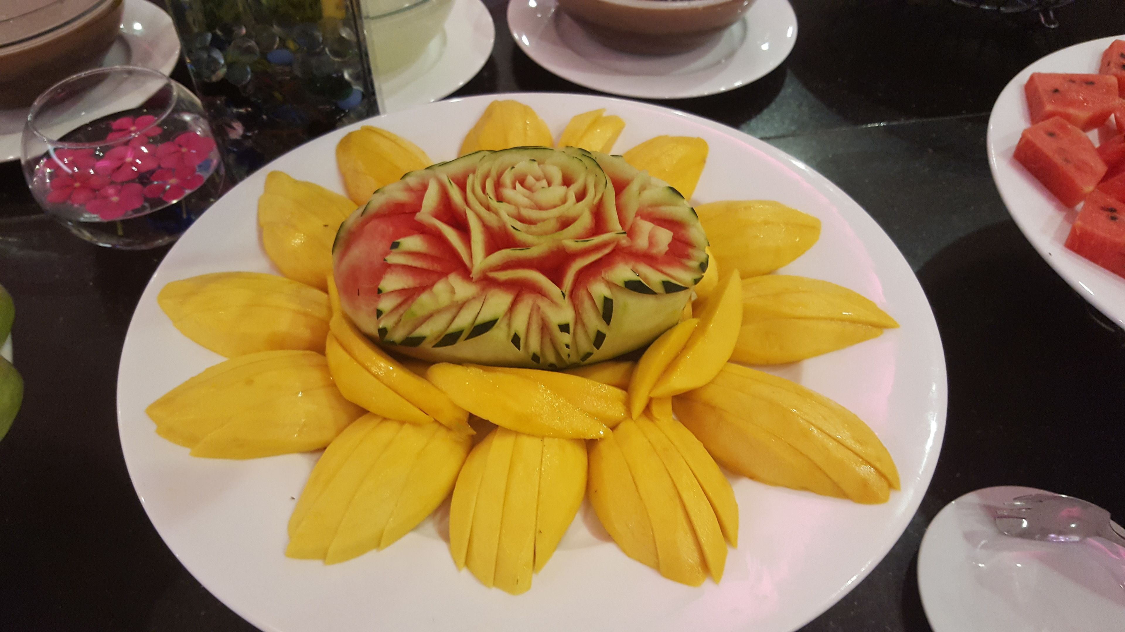 Une assiette ornée d’une sculpture de pastèque en forme de fleur, entourée de tranches de mangue.