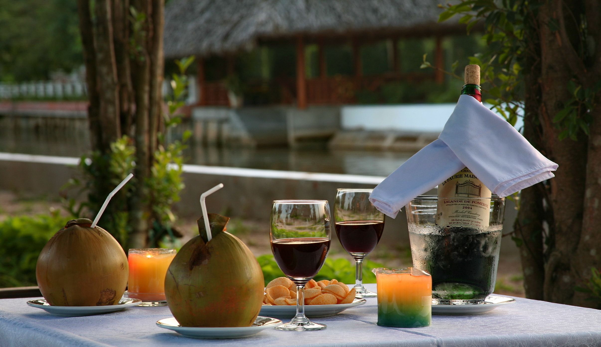 A table set with drinks, snacks, and a bottle of champagne, with a view of a thatched-roof building in the background.