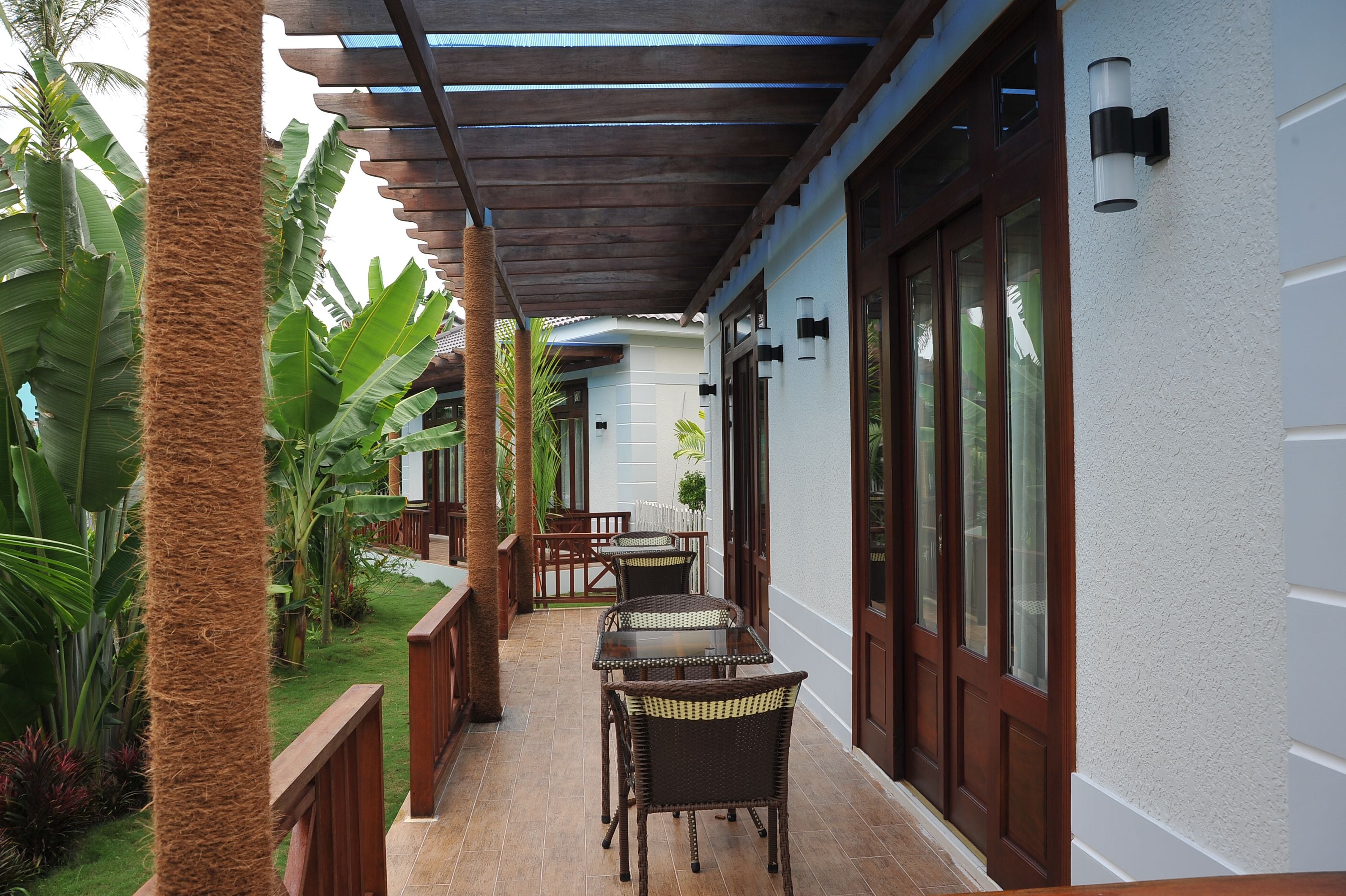A covered outdoor area with a table and chairs, surrounded by lush greenery and a white building with large windows.