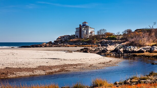 A coastal house with a prominent tower, situated on a rocky shoreline with a beach and a small stream.
