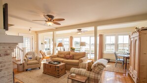 A spacious living room with a large sofa, armchairs, a coffee table, and a dining area with a table and chairs. There are ceiling fans, a fireplace, and large windows providing natural light.