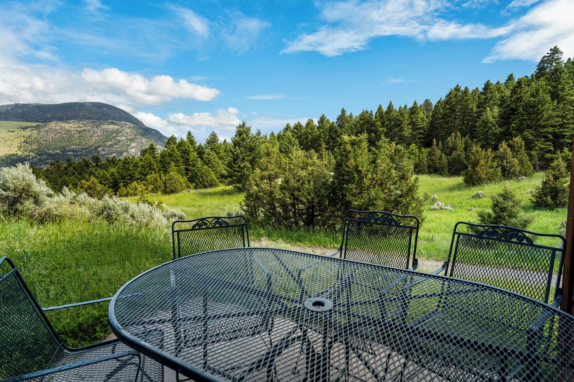 Une table et des chaises en métal sont disposées à l’extérieur, offrant une vue sur une forêt et des montagnes.