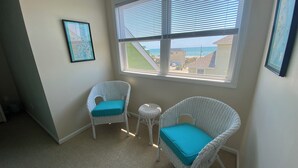 A corner room with two wicker chairs with teal cushions, a small round table, and a large window with blinds. - Panoramic Ocean & Sound Views, 2 min walk to beach, Sleeps 8-10, Emerald Isle (Emerald Isle)