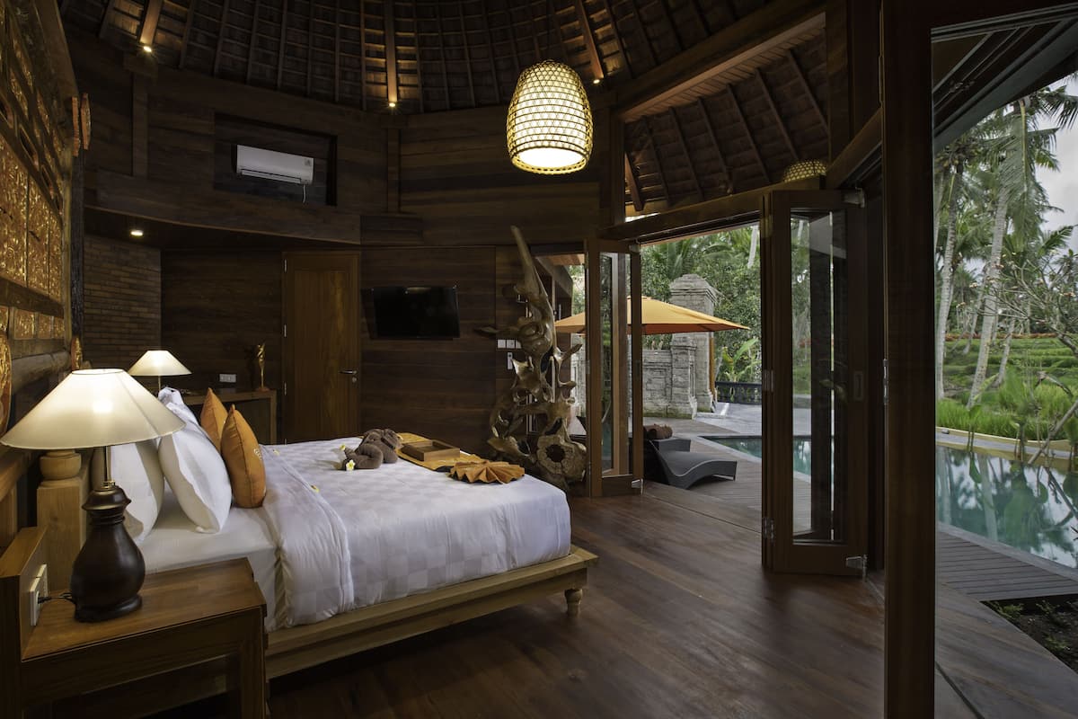 A spacious bedroom with a wooden bed, a nightstand with a lamp, and a large window offering a view of a pool and tropical vegetation.