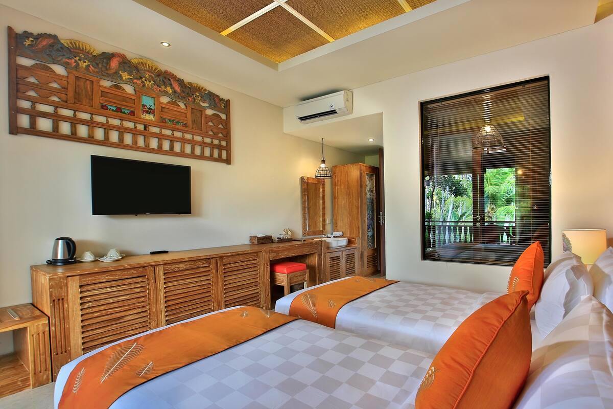 A hotel room with two beds, a wooden desk, a television, and a large window with blinds.