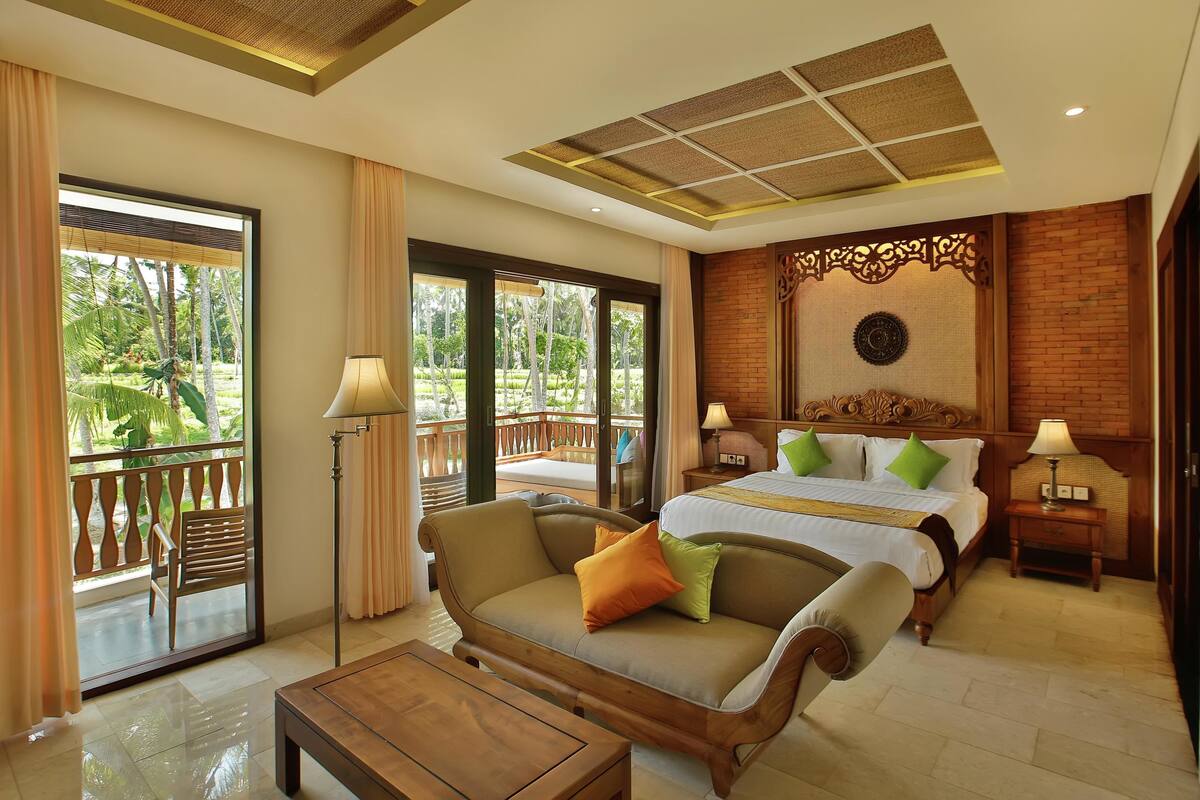 A hotel room with a bed, a sofa, a coffee table, and a balcony with a view of greenery.