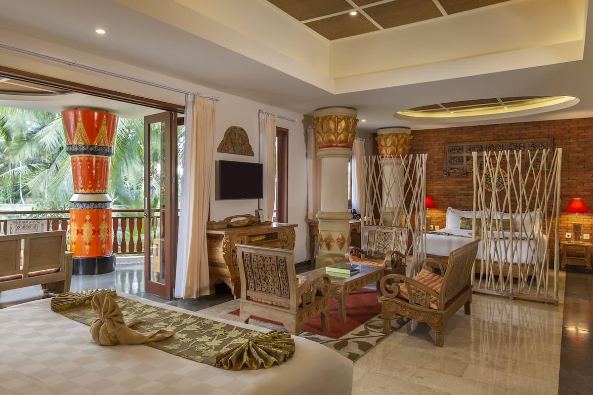 A spacious room with a large bed, a wooden desk, and a comfortable seating area. There is a balcony with a view of palm trees.