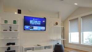 A modern living room with a flat-screen TV mounted on the wall, wooden flooring, white shelving units, and a large window with blinds. - Modern Studio Apartment., Sleeps 4, Steps to the Beach! (Santa Rosa Beach)