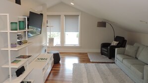 A modern living room with a high ceiling, wooden flooring, a grey sofa, a black leather chair, a white entertainment unit with shelves, and a large window with blinds. - Modern Studio Apartment., Sleeps 4, Steps to the Beach! (Santa Rosa Beach)