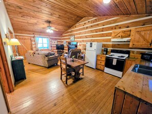 A wooden cabin interior with a living room, dining area, kitchen, and a ceiling fan.