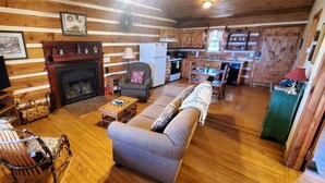 A cozy cabin interior with a fireplace, wooden walls, a sofa, a television, and a kitchen area. - Nite Vu Cabin:A Light and Airy Retreat with Mountain Views, Hot Tub, and Jacuzzi (Sevierville)