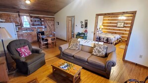 A cozy living room with a sofa, armchair, and coffee table. A kitchen area with wooden cabinets and a dining table is visible in the background. - Nite Vu Cabin:A Light and Airy Retreat with Mountain Views, Hot Tub, and Jacuzzi (Sevierville)