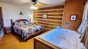A bedroom with a wooden floor, a bed with a patterned bedspread, a large bathtub, and a ceiling fan. - Nite Vu Cabin:A Light and Airy Retreat with Mountain Views, Hot Tub, and Jacuzzi (Sevierville)
