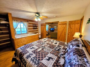 Ein Schlafzimmer in einer Blockhütte mit einem großen Bett, einem Fenster mit Vorhängen, einem Deckenventilator und einem Fernseher in der Ecke.