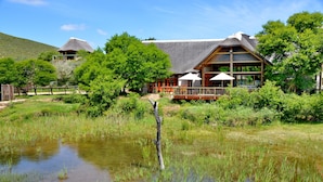 Een lodge met rieten dak, een houten terras en grote ramen met uitzicht op een vijver.