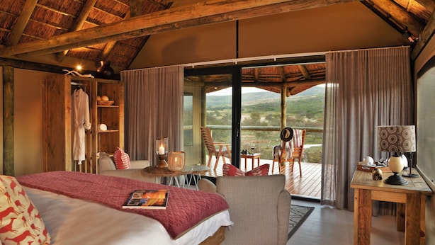 Een slaapkamer met een groot bed, een houten plafond, een balkon met een tafel en stoelen, en uitzicht op het landschap.
