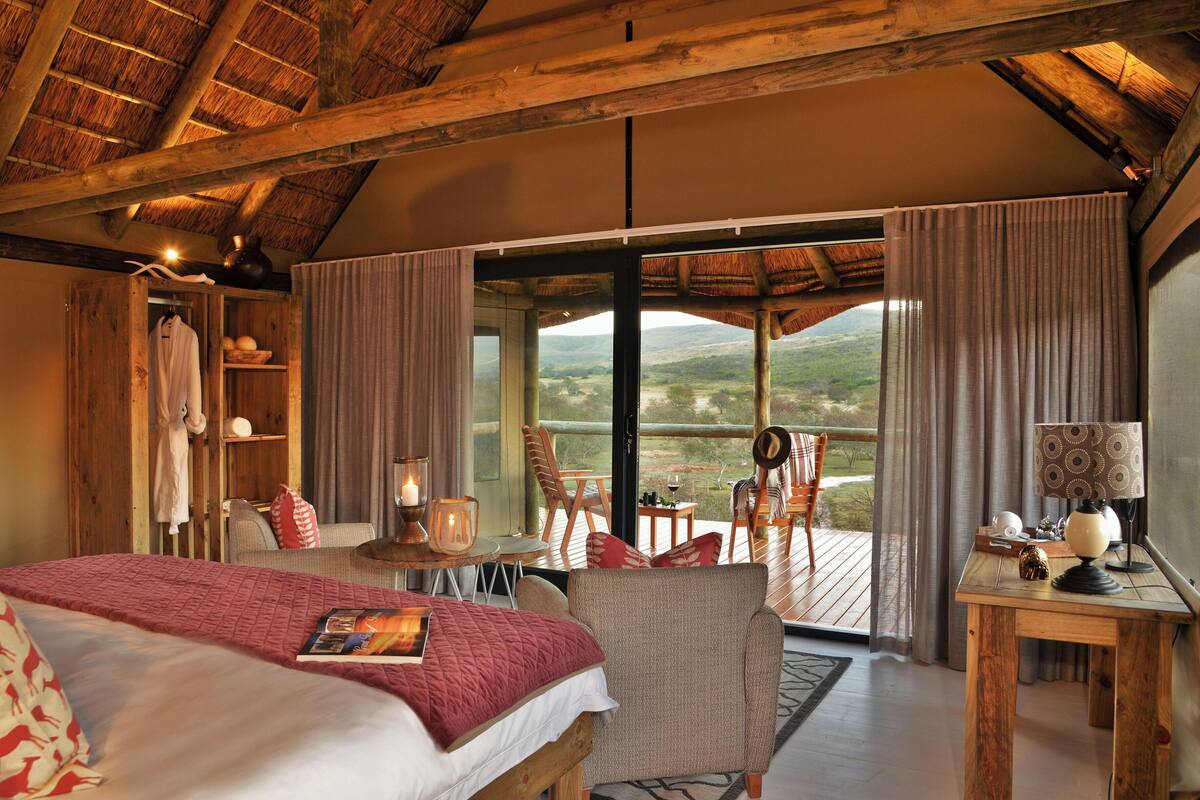 Une chambre avec un grand lit, un plafond en bois, un balcon avec une table et des chaises, et une vue sur la campagne.