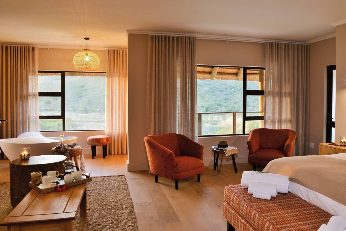 Une chambre d’hôtel avec un grand lit, deux fauteuils, une petite table, une baignoire et de grandes fenêtres avec des rideaux.