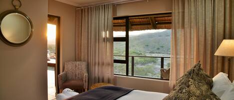A hotel room with a bed, a chair, a mirror, a window with a view of the countryside, and a lamp.