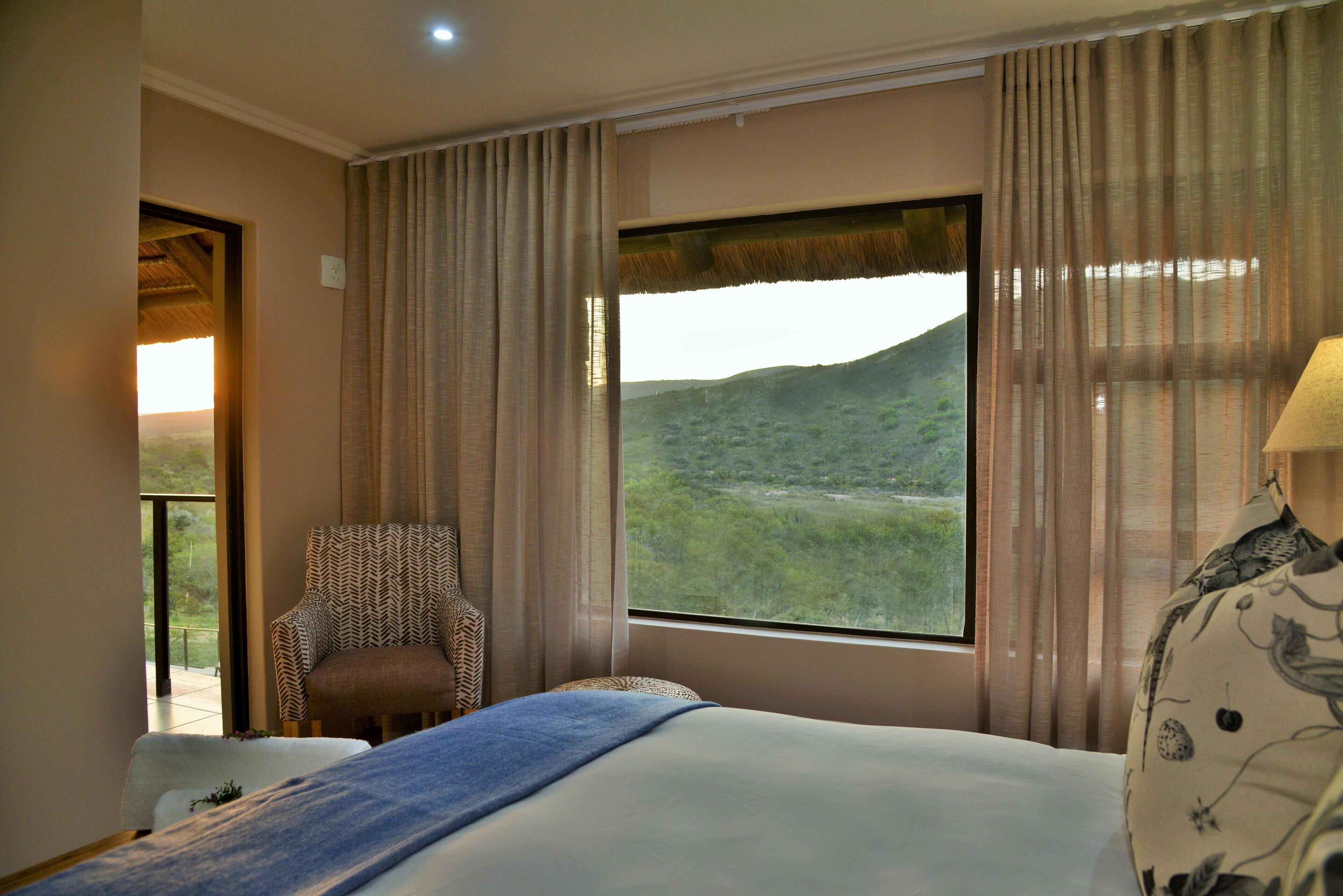A hotel room with a large bed, a chair, a window with a view of a hilly landscape, and a lamp on the right side.