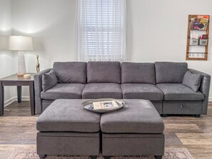 Ein graues Ecksofa mit Serviertablett davor, ein kleiner Tisch mit einer Lampe und eine gerahmte Wandtafel mit Spielboxen.