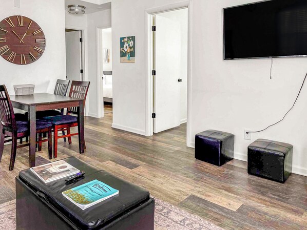 A living room with a dining area, a clock, a flat-screen TV, and a small black speaker.