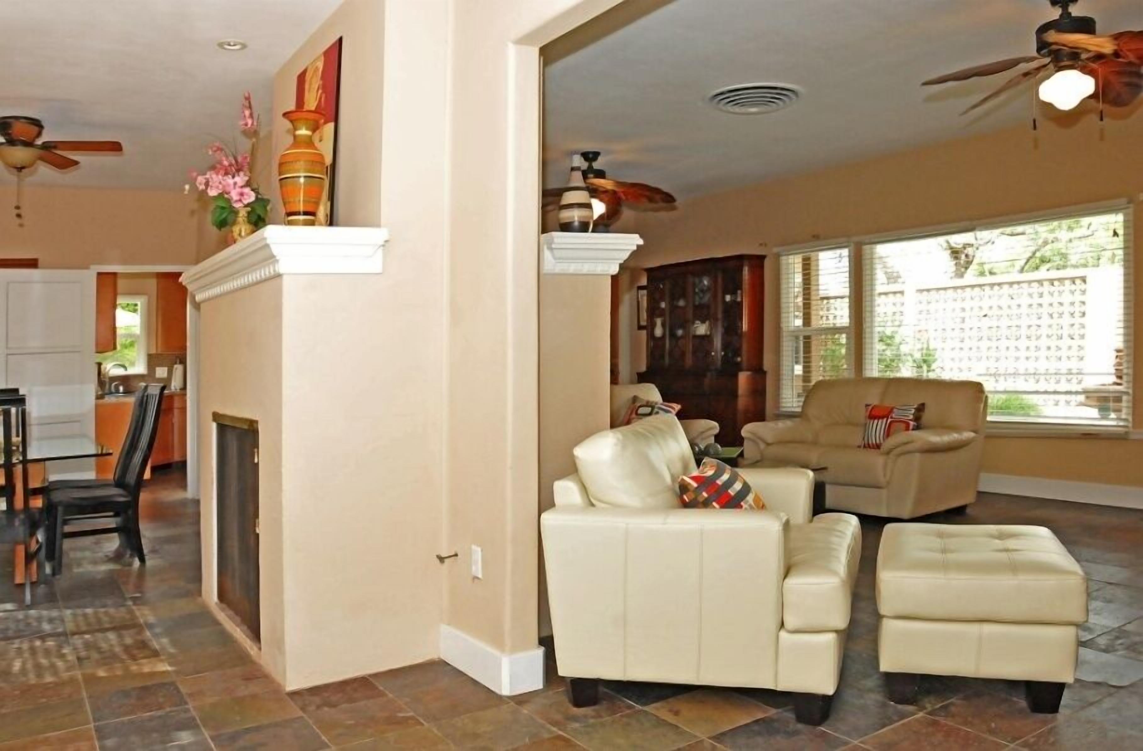A spacious living room with a fireplace, a ceiling fan, and a large window with blinds.