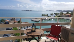 Ein Balkon mit Tisch und Stuhl, mit Blick auf einen Yachthafen mit Booten und einer entfernten Insel.