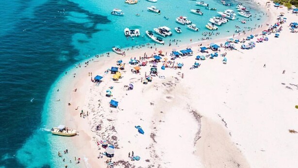 Ein Luftbild einer Strandbucht mit zahlreichen Booten, die im Wasser vor Anker liegen, einem sandigen Ufer mit Strandsonnenschirmen und einem strahlend blauen Himmel.