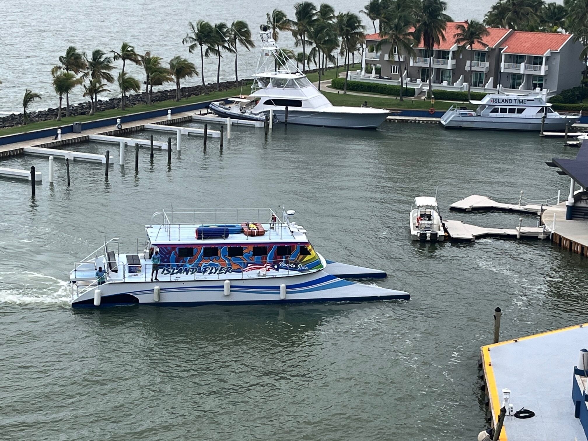 A ferry boat with "Island Flyer" written on the side, docked at a marina with palm trees and a building in the background.