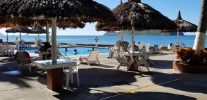 A poolside area with thatched umbrellas, lounge chairs, and a view of the sea. - Spectacular Corner Unit w/Huge Ocean Views, Pool & Private Beach Access (Mazatlan)
