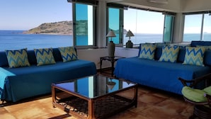 A room with two blue sofas, a glass coffee table, and a green chair. Large windows offer a view of the sea and a distant island.