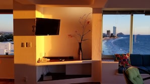 A living room with a TV mounted on the wall, a vase with flowers, a sofa with colorful cushions, and a large window overlooking a beach and buildings.