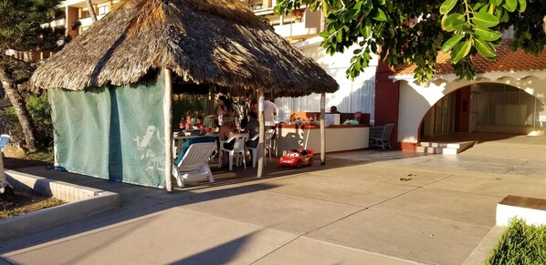 A thatched-roof outdoor seating area with plastic chairs and a table, surrounded by a building with balconies. - Spectacular Corner Unit w/Huge Ocean Views, Pool & Private Beach Access (Mazatlan)