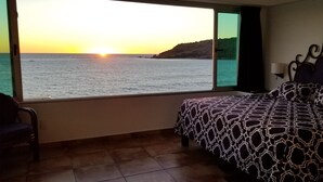 A bedroom with a bed, a chair, and a large window offering a view of the sea and a sunset.