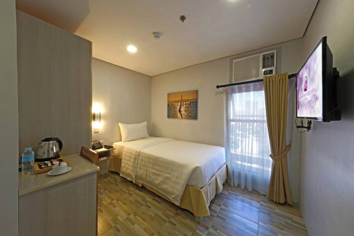A hotel room with a bed, a desk with a kettle, a phone, and a painting on the wall.