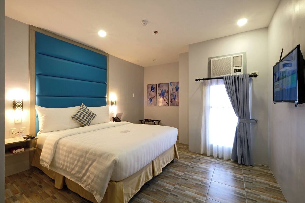 A hotel room with a large bed, bedside tables, a flat-screen TV, a window with curtains, and artwork on the wall.