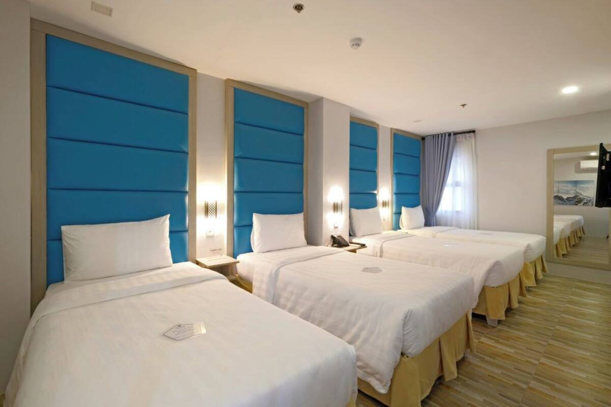 A hotel room with four single beds, each with a white bedspread and a headboard with a blue panel. There are bedside tables with lamps and a mirror on the wall.