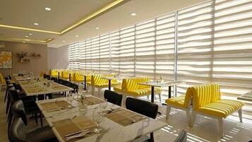 Un comedor con mesas dispuestas para comer, sillas amarillas y ventanas grandes con persianas.