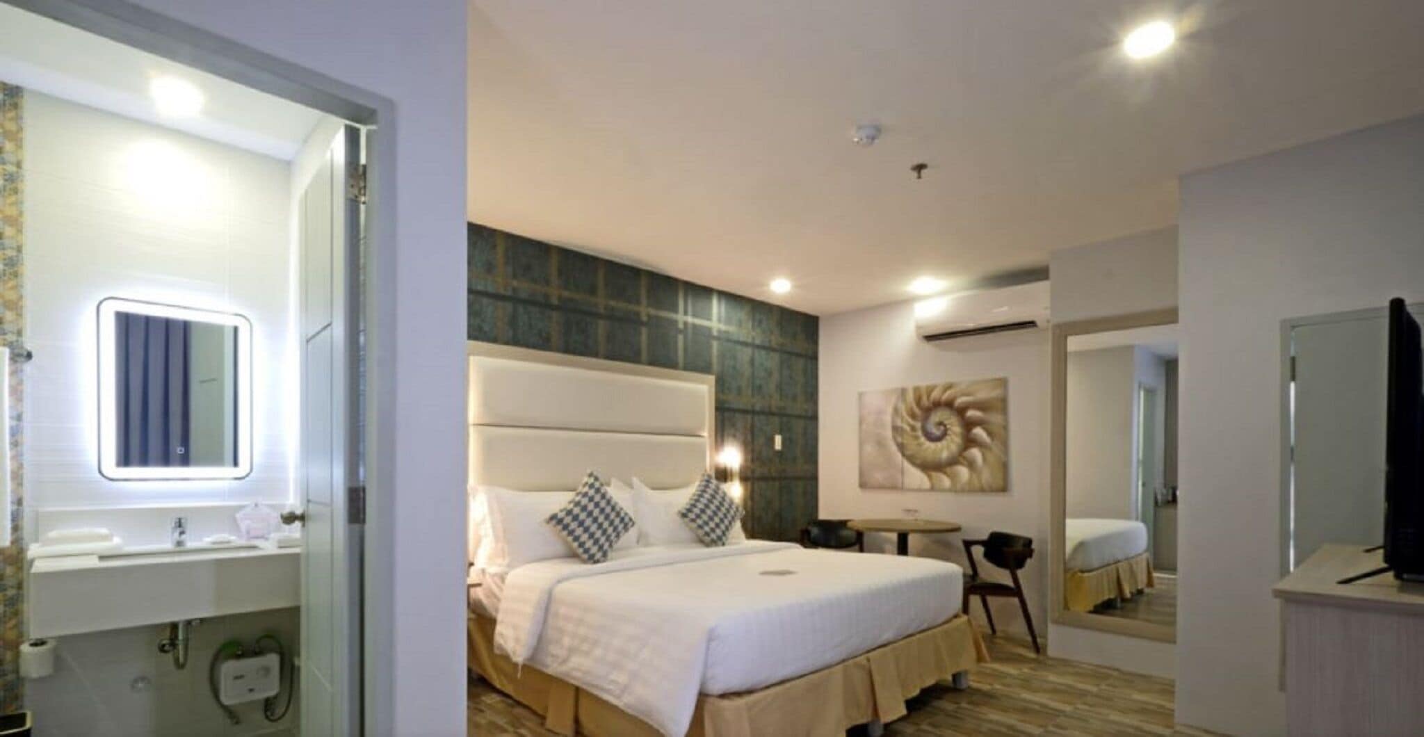 A hotel room with a bed, bedside table, mirror, and a painting on the wall.