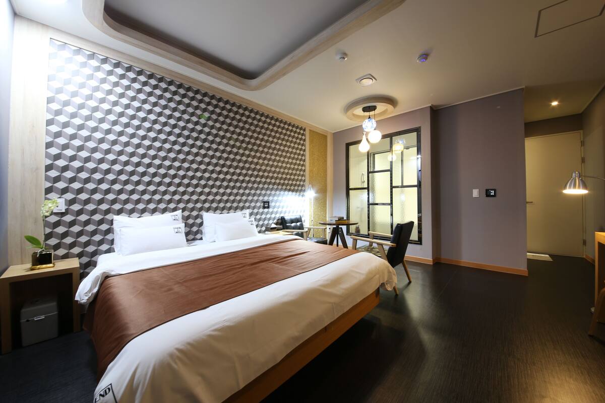 A modern hotel room with a large bed, a geometric patterned wall, a small table with chairs, and a large window.