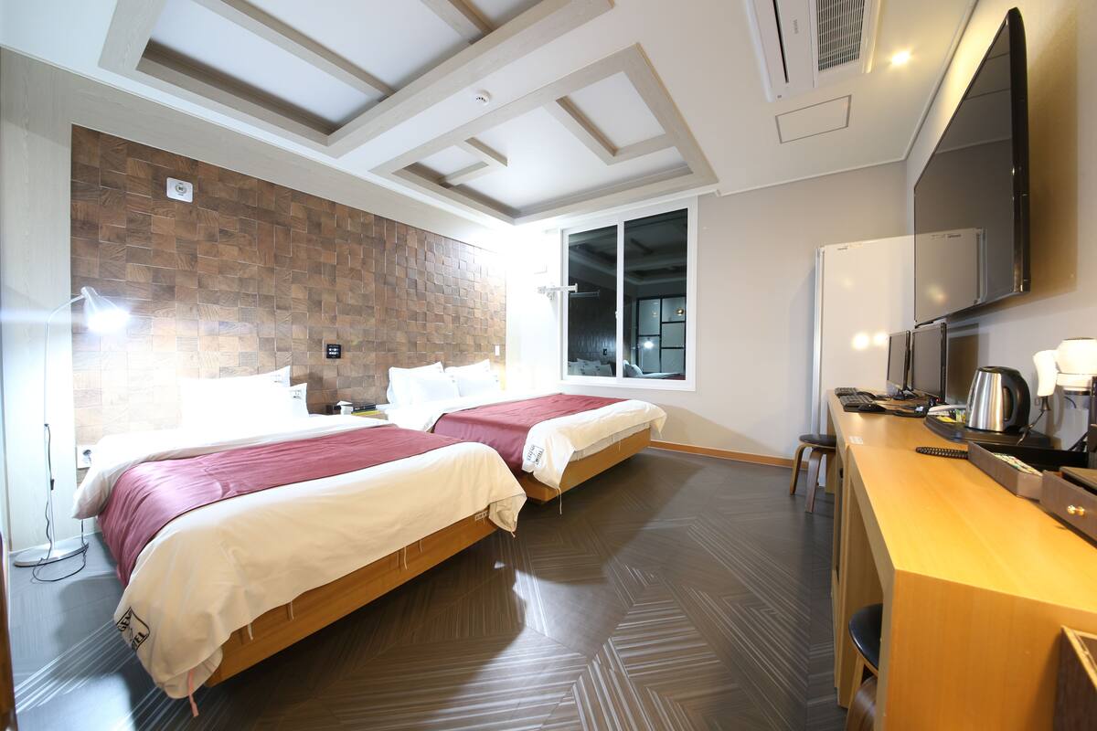 A hotel room with two beds, a desk with a computer, a flat-screen TV, and a large window.