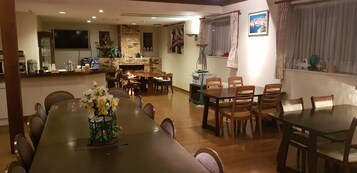 Un vaste espace dédié à la salle à manger, avec une longue table et de nombreuses chaises, un coin cuisine doté d’une cheminée en pierre, et une fenêtre agrémentée de rideaux.