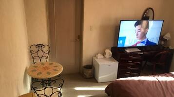 A hotel room with a bed, a desk with a computer, a television, a small table with a chair, and a door.
