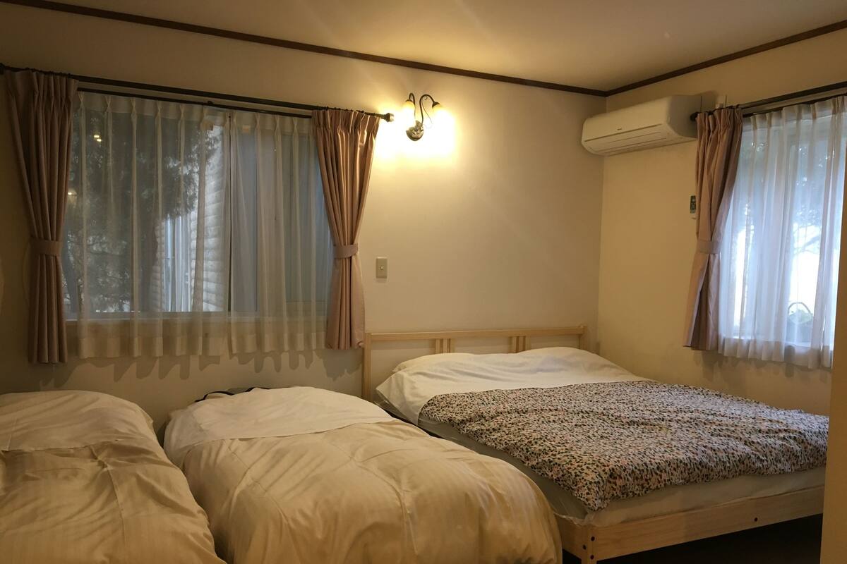 A room with two single beds, a wooden headboard, beige bedding, sheer curtains, and an air conditioning unit.