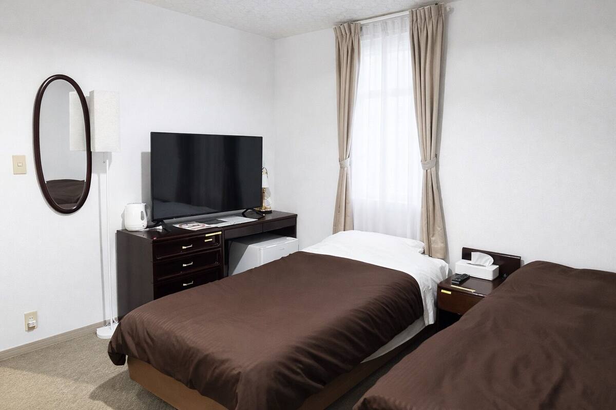A hotel room with two beds, a desk with a flat-screen TV, a mirror, and a window with curtains.