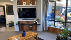 Un salon moderne doté d’un grand téléviseur à écran plat fixé au mur, d’une table basse en bois avec des télécommandes, d’un canapé en cuir noir et d’une grande fenêtre donnant sur la rue.