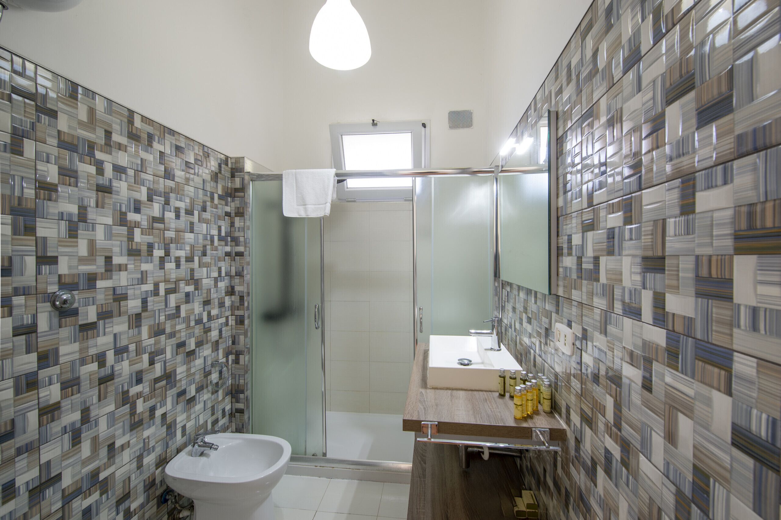 A bathroom with a tiled wall, a sink, a mirror, and a shower.