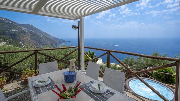A terrace with a table set for dining, a hot tub, and a scenic view of the sea and mountains.