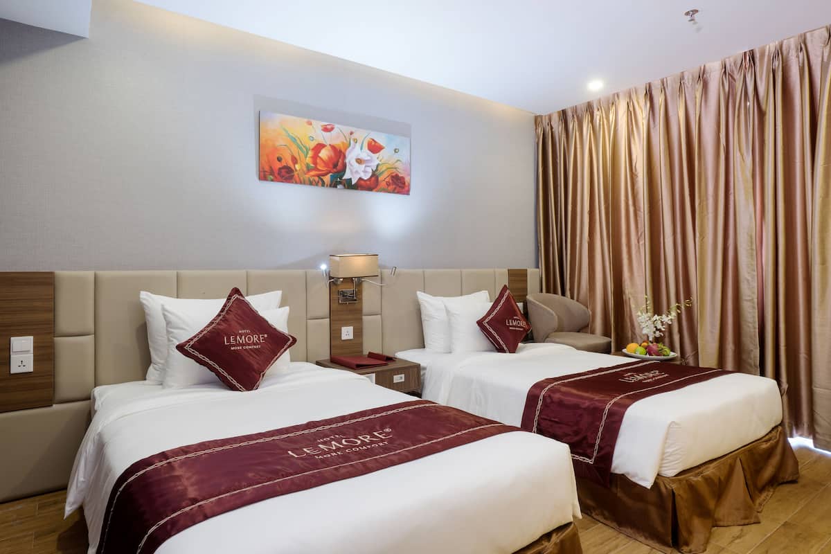 A hotel room with two beds, each with a maroon bedspread and matching pillows, a nightstand with a lamp, a painting on the wall, and a large window with curtains.